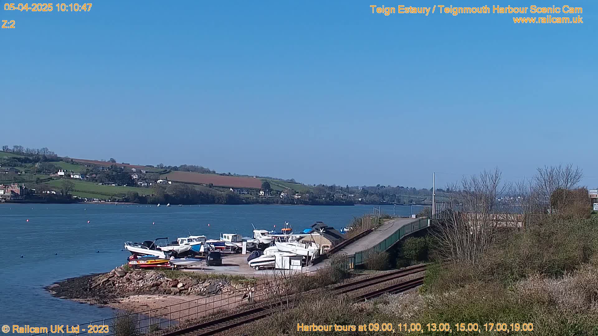Several small boats are moored at a harbor next to a railway line under a clear blue sky.