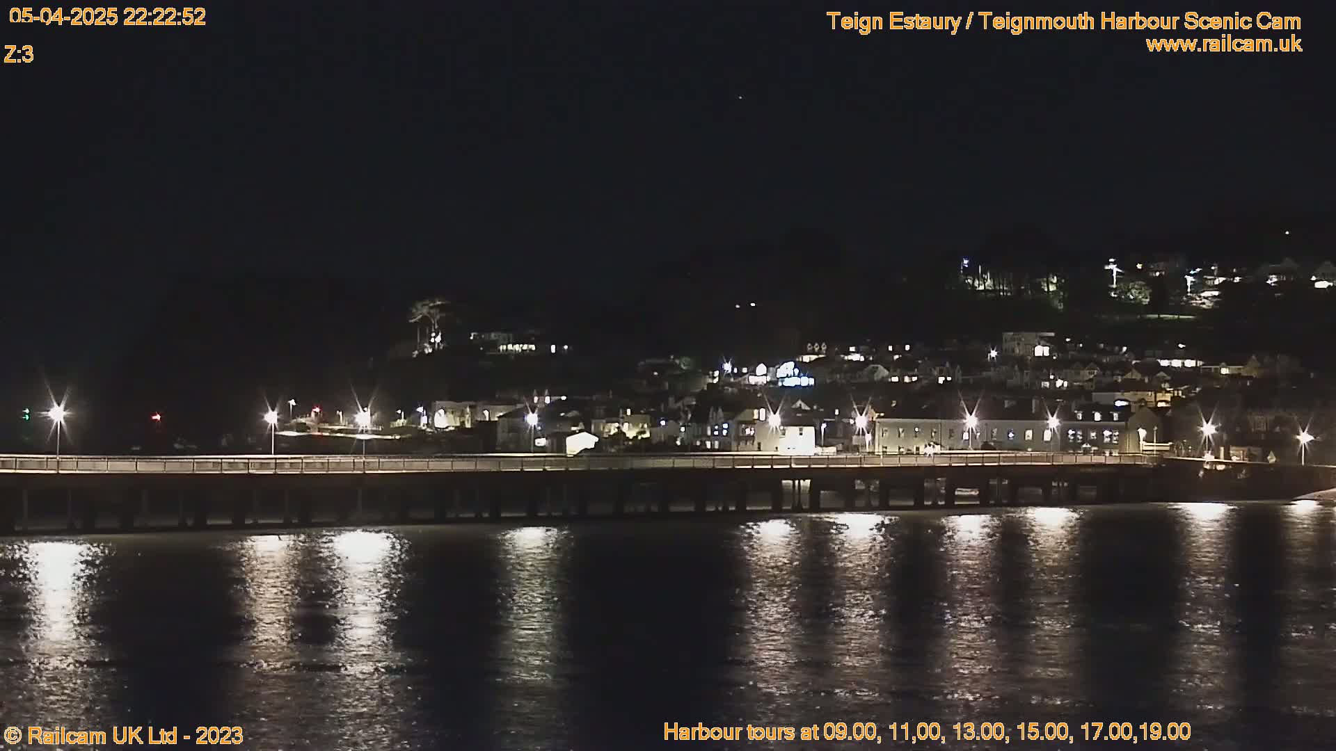 A nighttime view of a pier extending into a calm body of water, with a town's lights illuminating the hillside in the background.