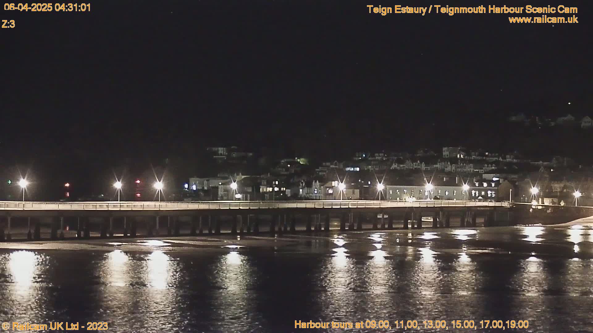 A long pier is illuminated at night, extending over calm, dark water reflecting the lights, with a town visible in the background under a clear, dark sky.