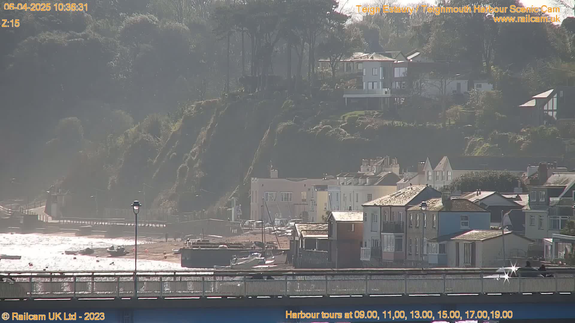 A hazy, sunny day reveals a coastal town nestled against a hillside, viewed from a bridge overlooking a calm harbor with several small boats.