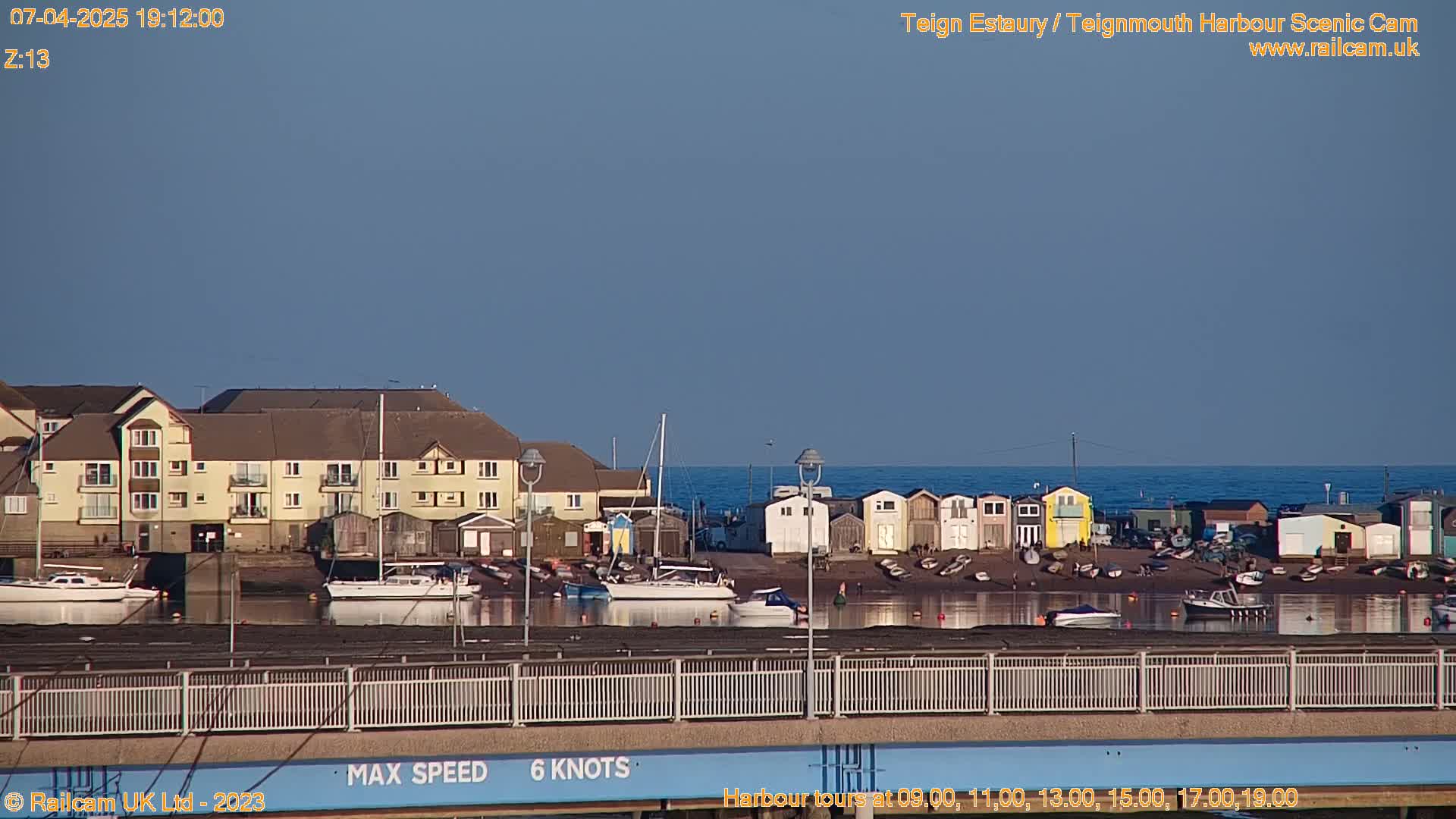 A calm harbor scene under a clear blue sky shows a row of colorful beach huts, boats docked in the water, and a multi-story apartment building in the background.
