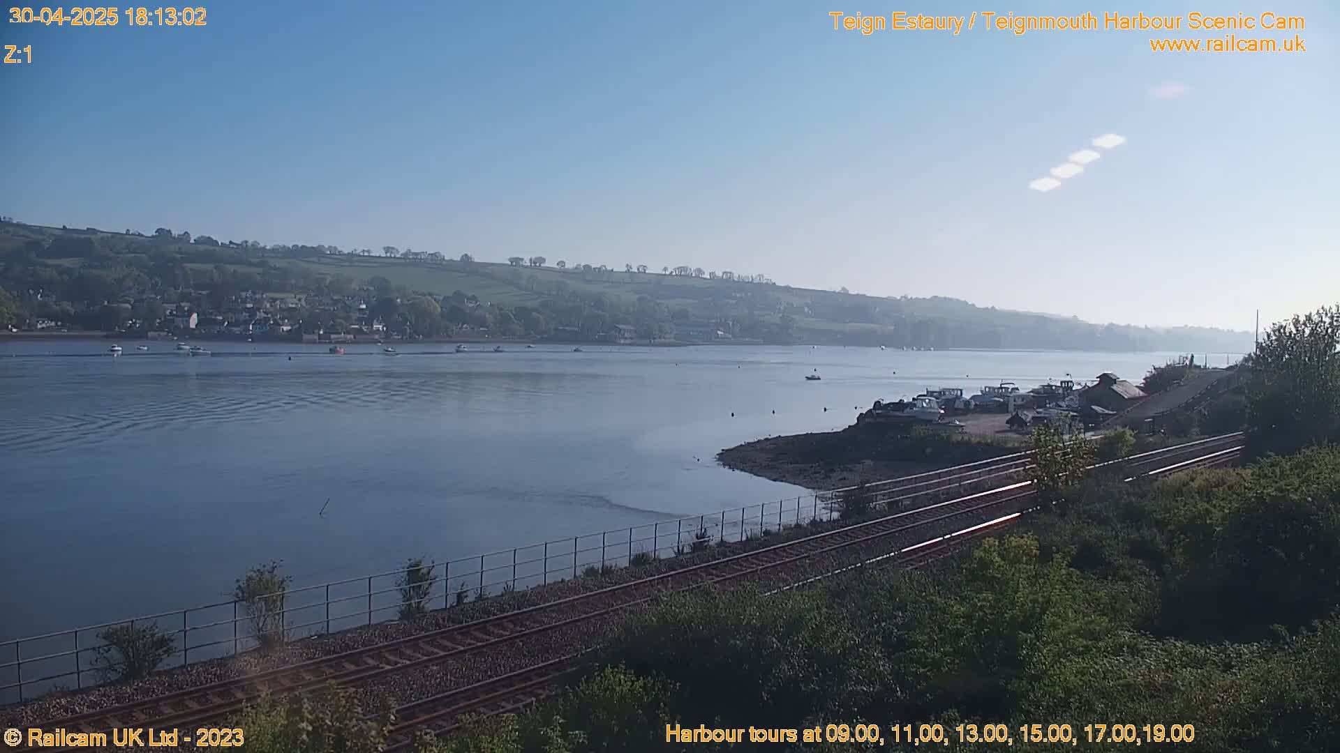 A calm, clear day shows a wide estuary with several boats, a small town on the far shore, and a railway line in the foreground.