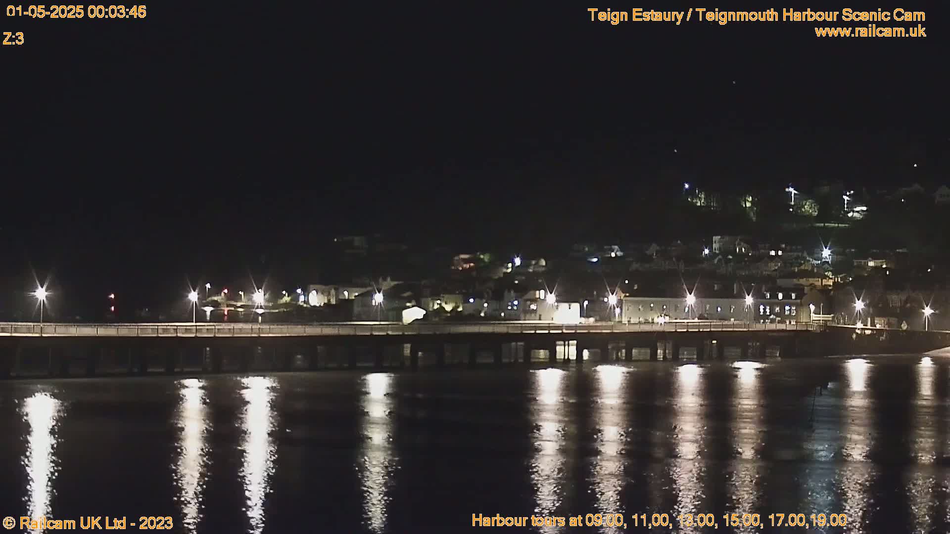 A long pier illuminated at night reflects in calm, dark water under a clear, starlit sky.