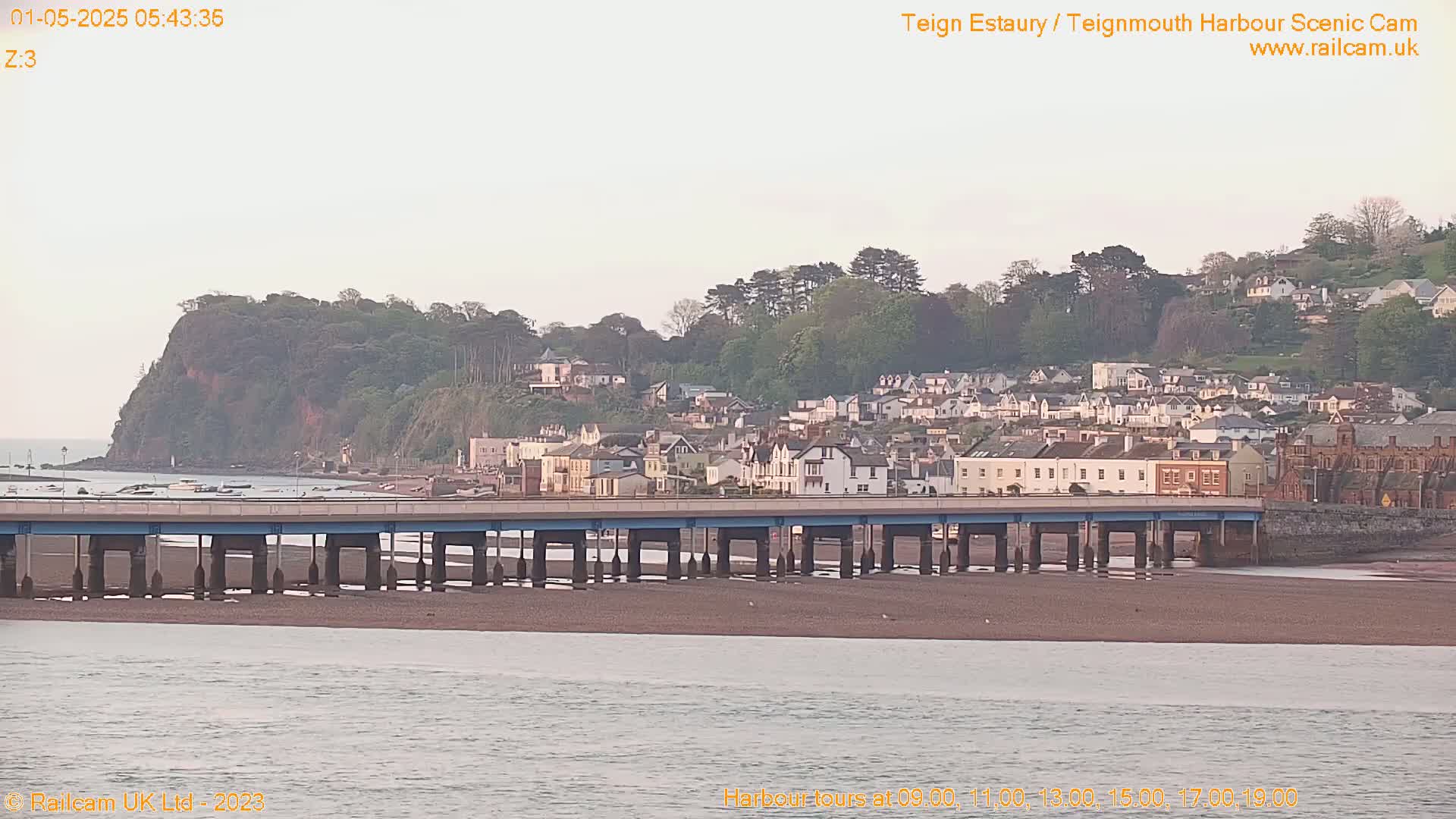A long bridge stretches across a calm body of water toward a town nestled on a hillside beneath a pale sky.