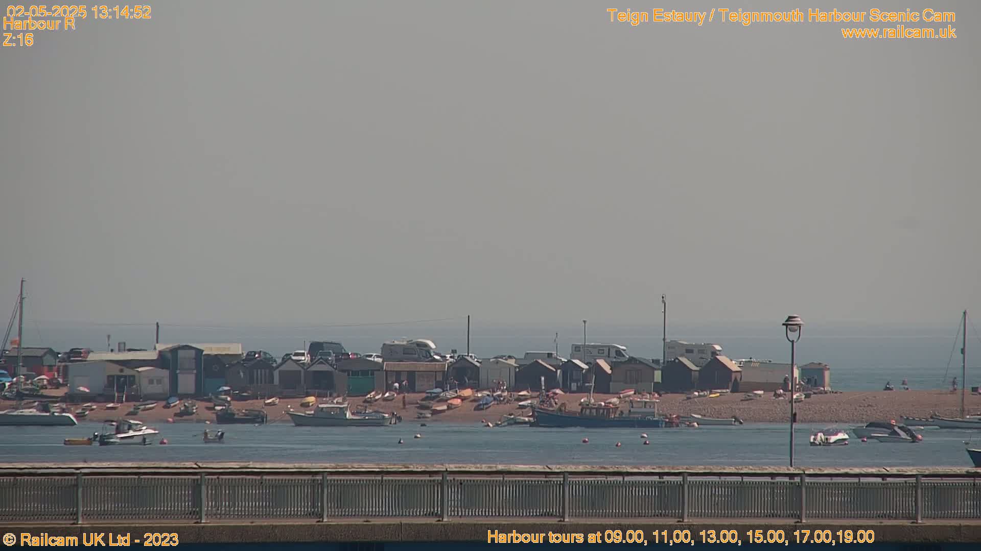 A hazy, sunny day reveals a beach with small buildings and numerous boats in a calm body of water, viewed from a distance across a railing.