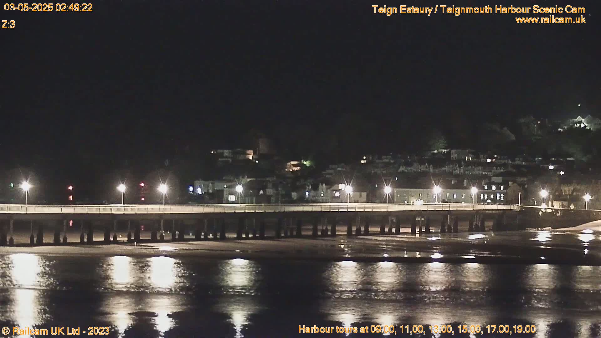 A long pier is lit at night, extending over a dark, calm body of water reflecting the lights, with a town visible in the background under a clear, dark sky.