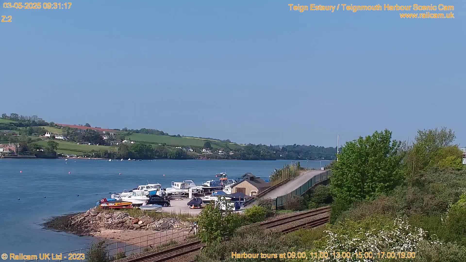 A sunny day shows a harbor with several boats docked near a small building and train tracks, beside a calm body of water with green hills in the distance.