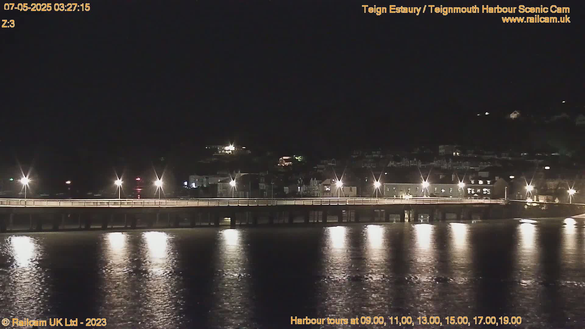 A long pier with lights stretches across calm, dark water at night, illuminated buildings visible in the distance.