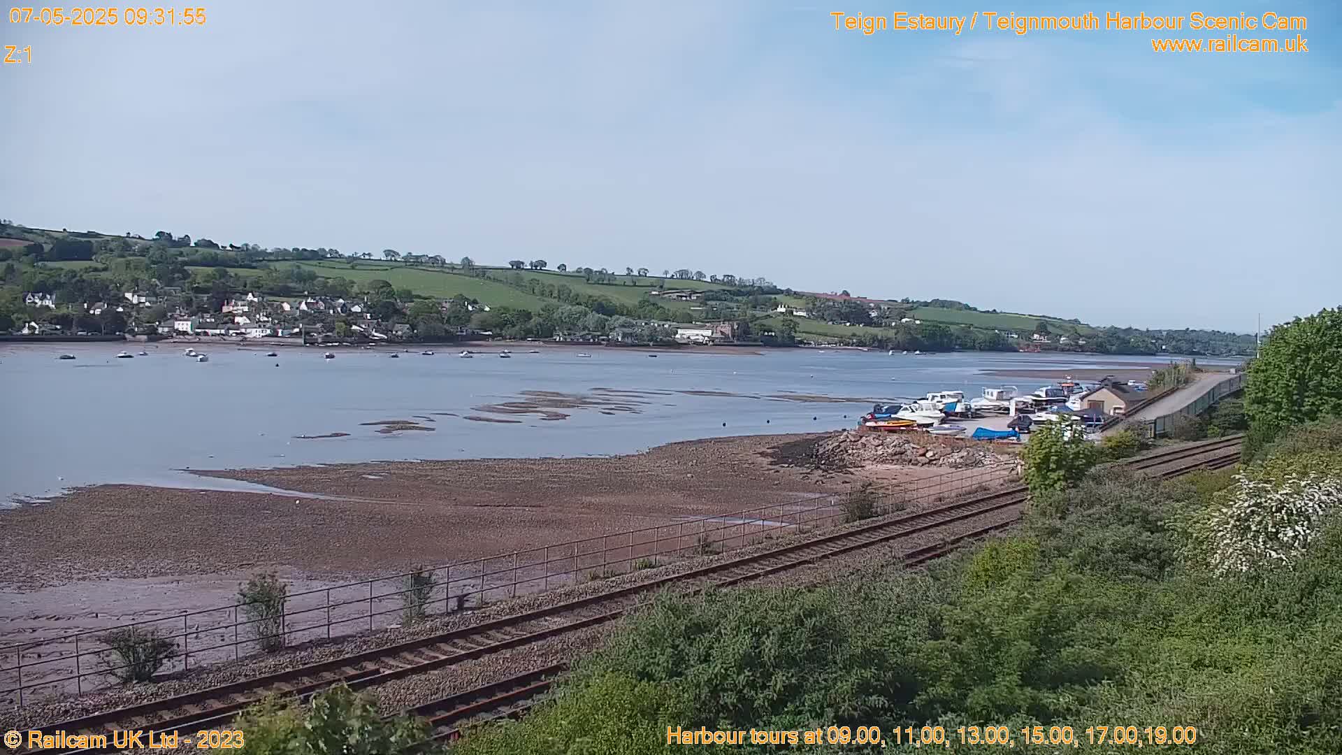 A calm, sunny day reveals a wide estuary with several boats moored near a railway line, and a small town nestled on a hillside in the distance.