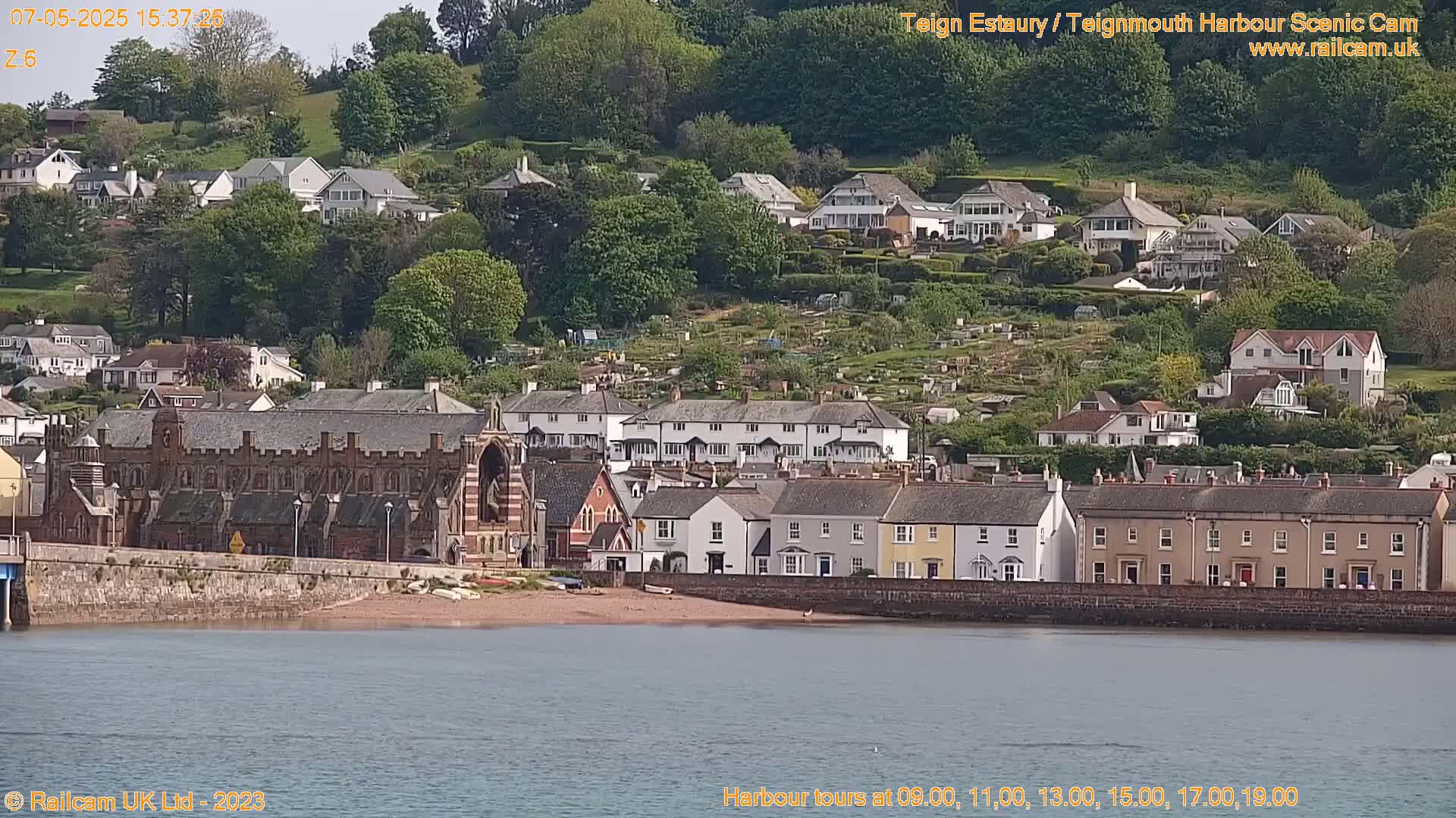 A seaside town with colorful houses and a large brick building sits on a hill overlooking calm water under a clear sky.