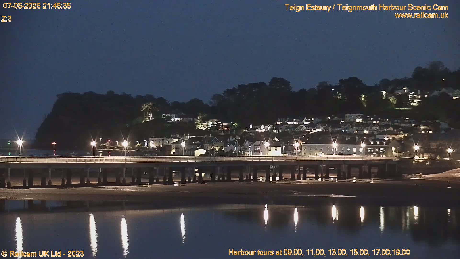 A long pier stretches across a calm, dark body of water at night, illuminated by lights lining the pier and reflecting in the water, with a hillside town visible in the background under a dark sky.