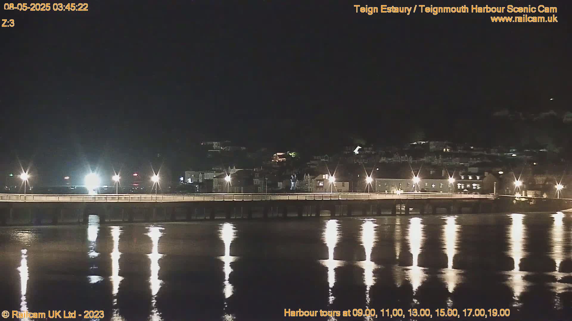 A nighttime view of a bridge lit by streetlights, reflecting in calm water under a dark sky.