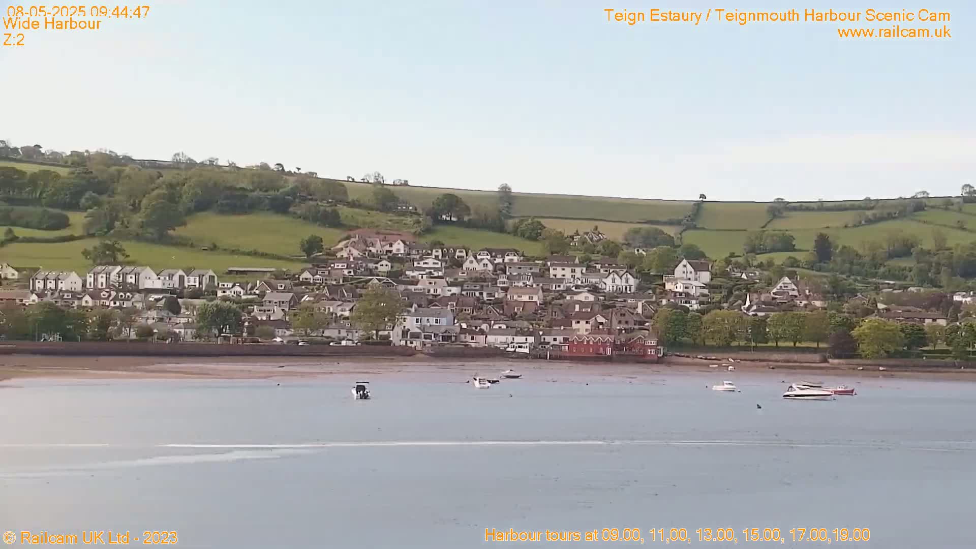 A coastal town with many houses sits on a gently sloping hillside overlooking a calm harbor with several small boats.