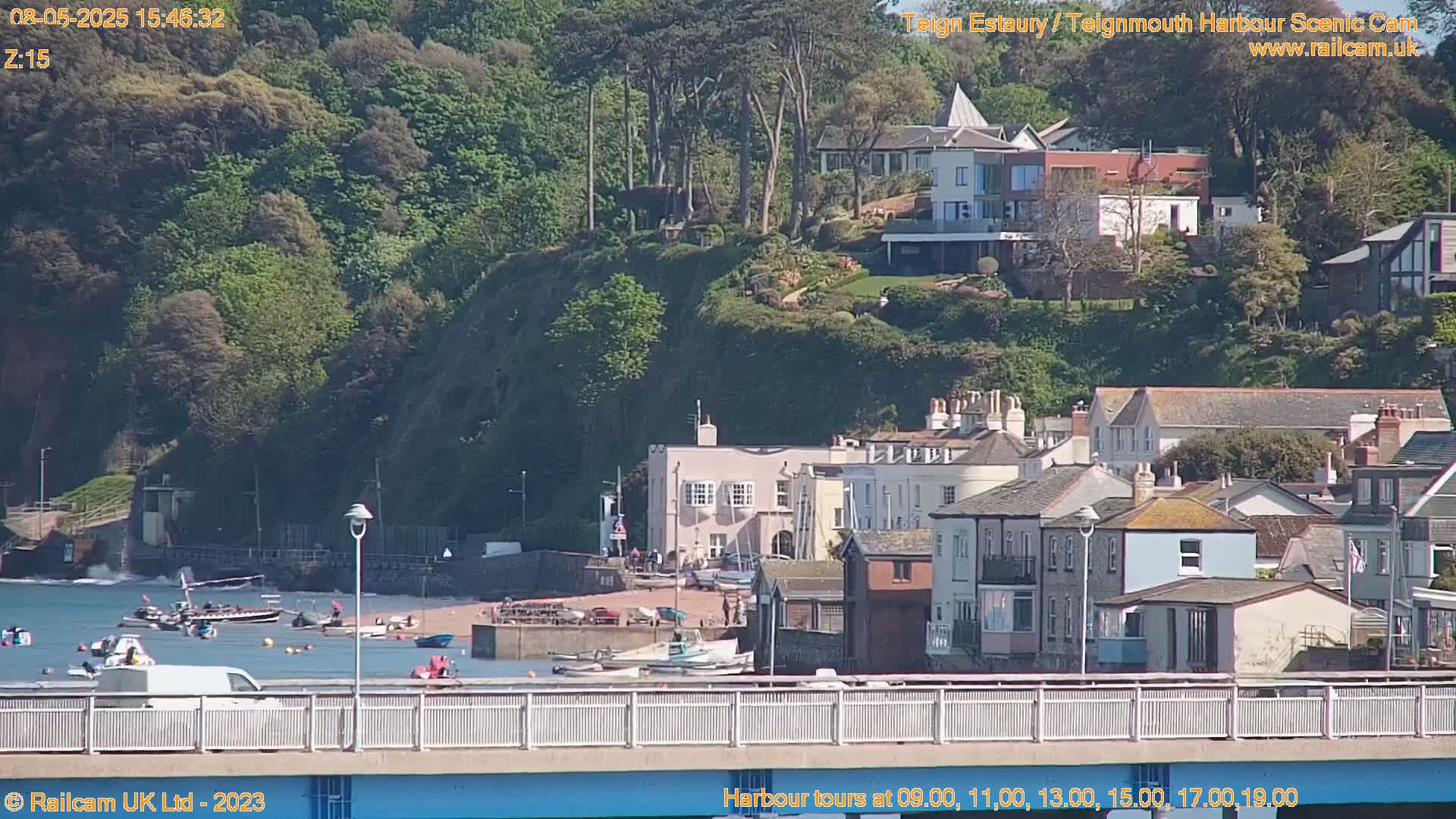 A sunny day shows a harbor scene with several boats on the water, a sandy beach with people, and a collection of buildings nestled on a hillside.