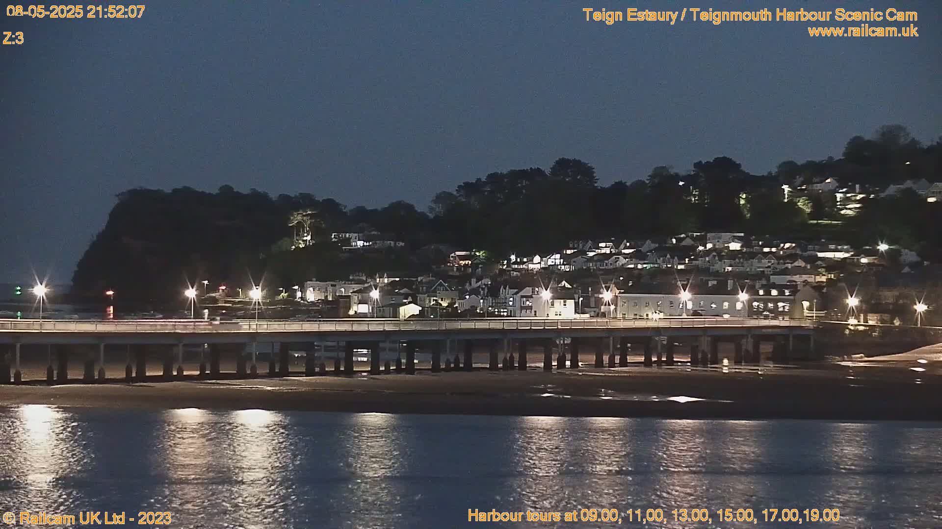 A nighttime view of a town nestled on a hillside overlooking a calm body of water, illuminated by lights along a pier and on the buildings.