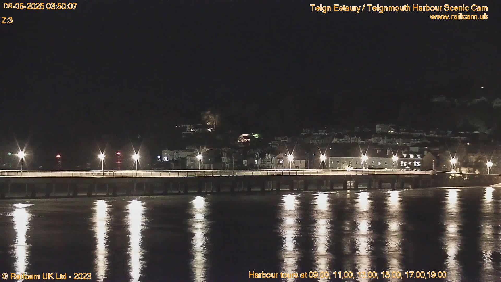 A long pier with lights is illuminated at night over calm, dark water reflecting the lights, with a town visible in the background.