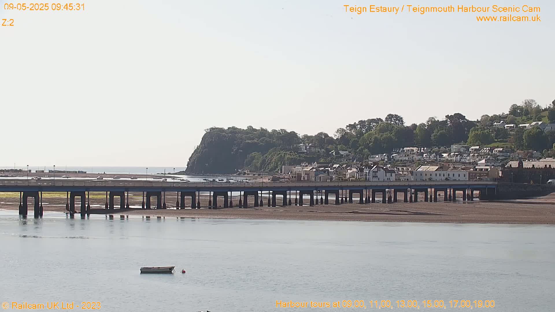 A long bridge stretches across a calm, wide waterway toward a town nestled against a green hill under a bright, clear sky.