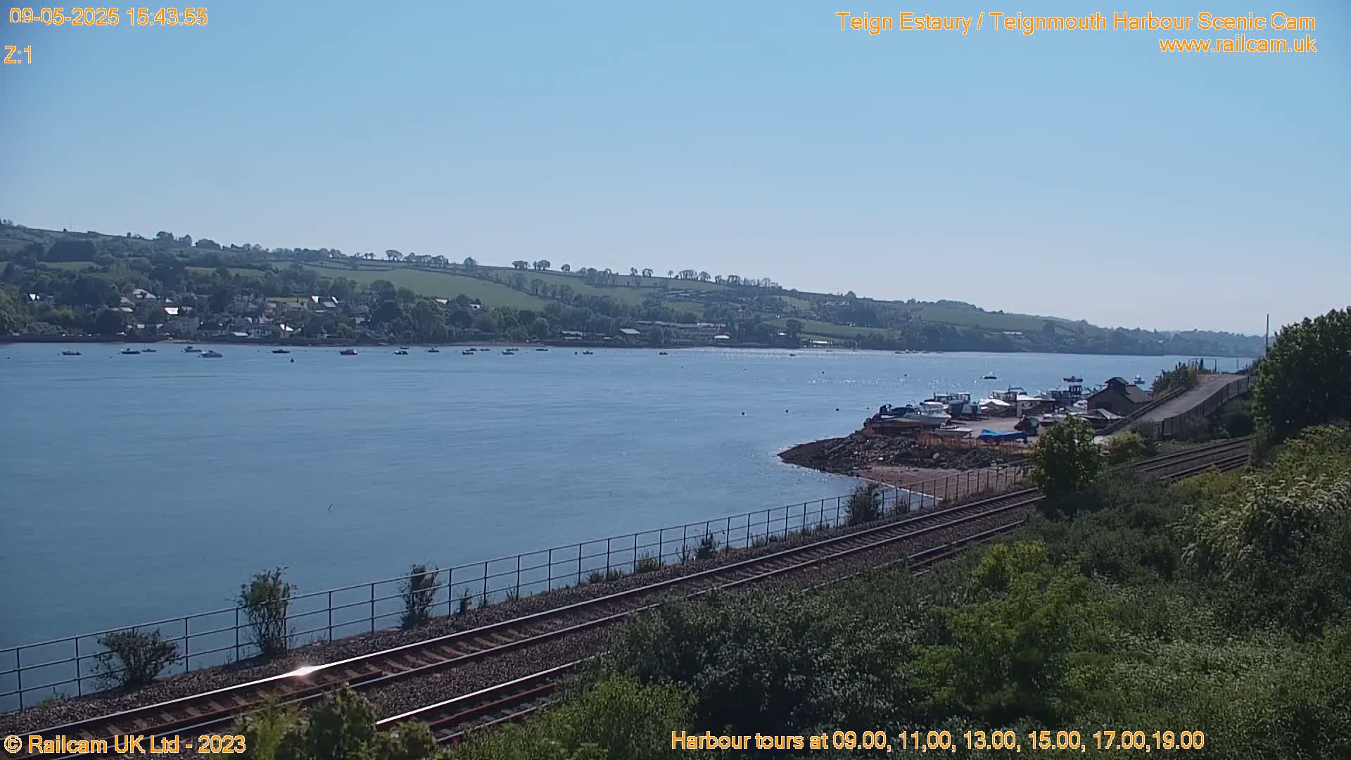 A calm body of water with several boats, a shoreline with a small harbor, and a railway track running alongside lush green vegetation under a clear blue sky.