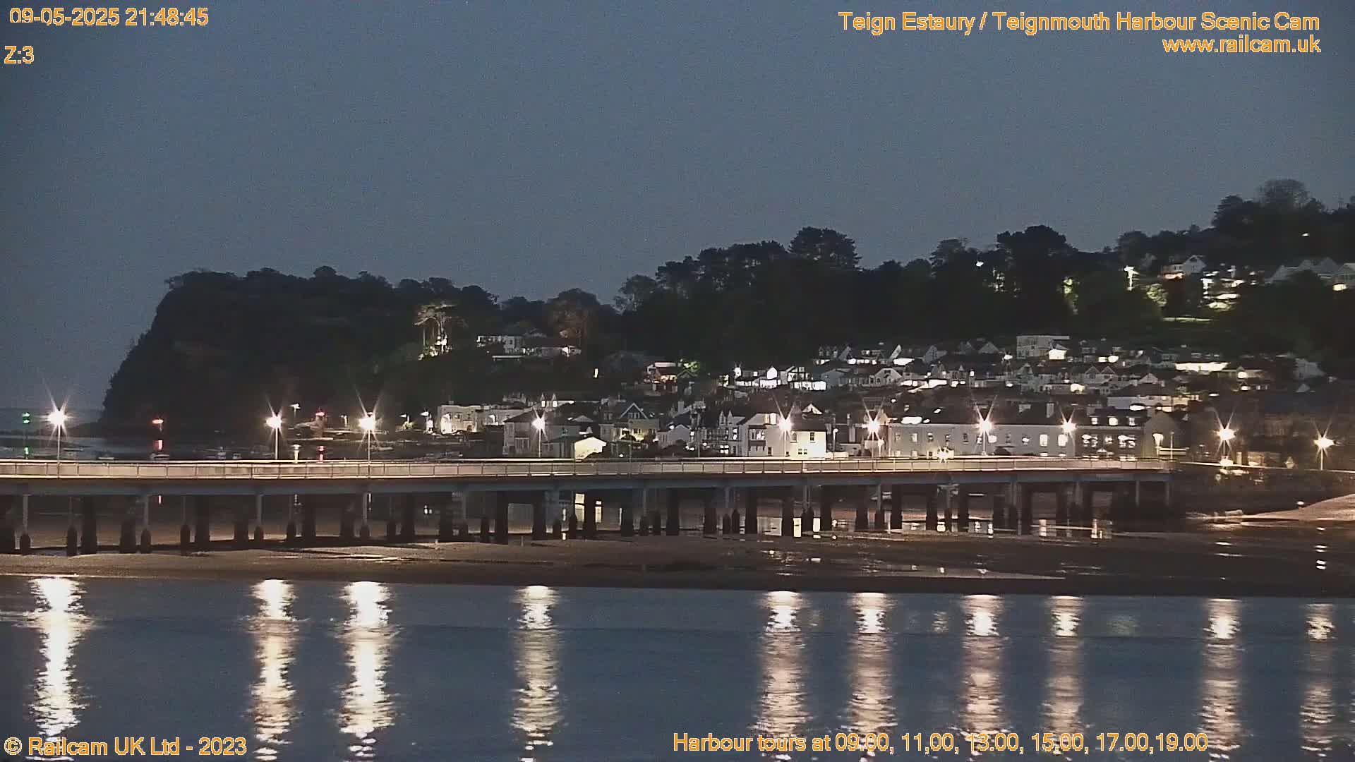 A long pier stretches across a calm, dark body of water reflecting lights from a nearby town nestled on a hillside under a twilight sky.