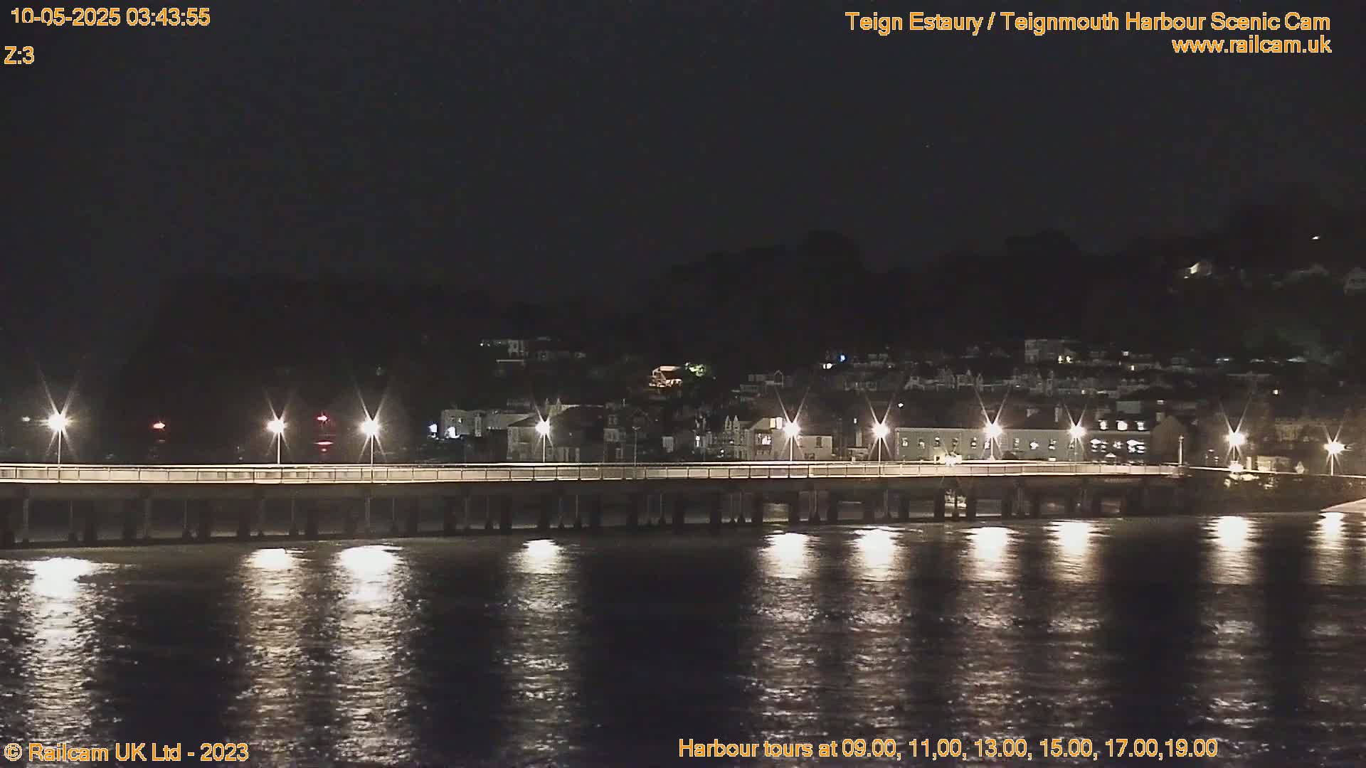 A long pier stretches across a dark, calm body of water at night, illuminated by lights along the pier and reflecting in the water, with a town visible in the background.