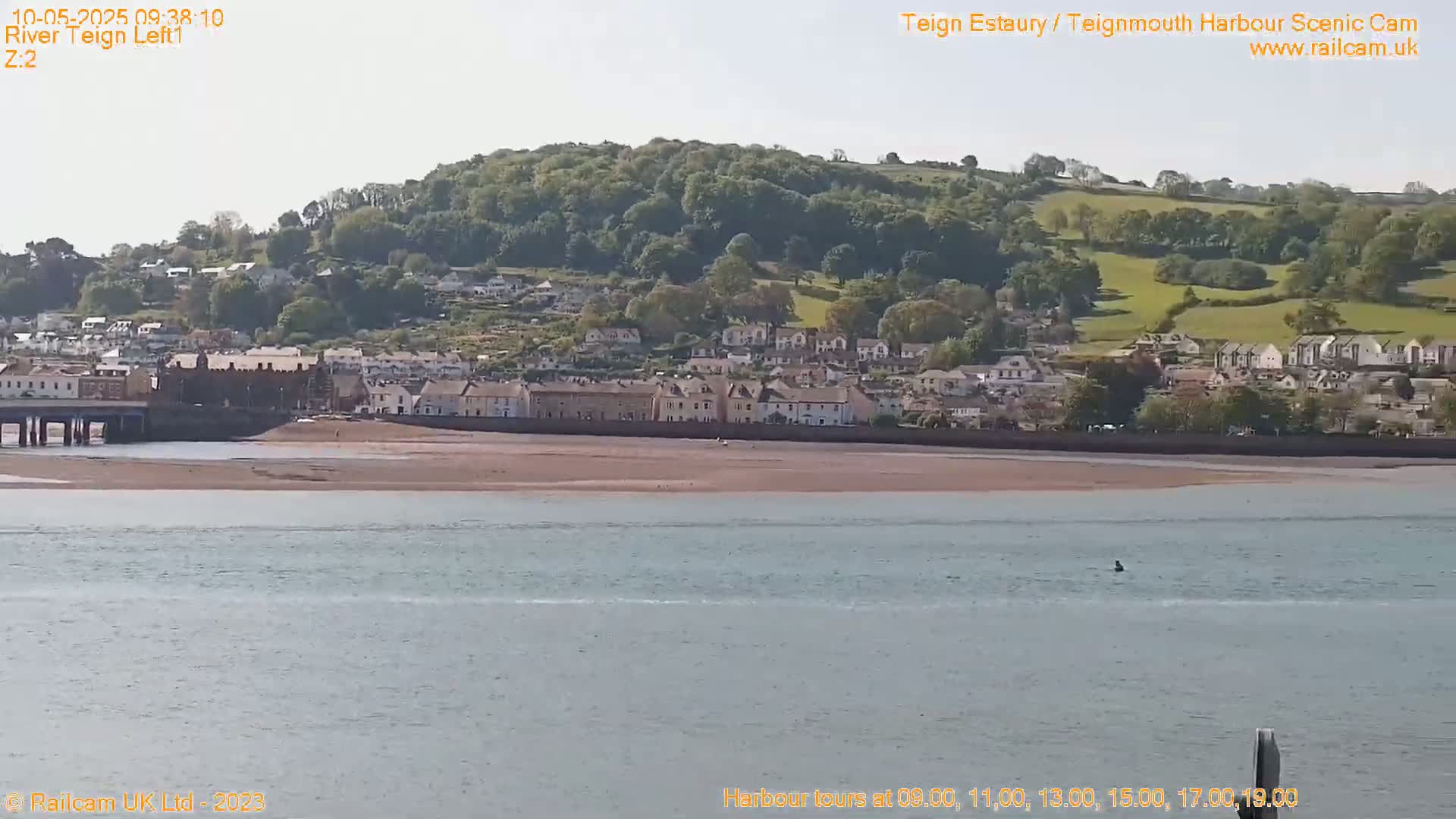 A seaside town nestled against a verdant hillside is visible across a calm body of water under a bright, sunny sky.
