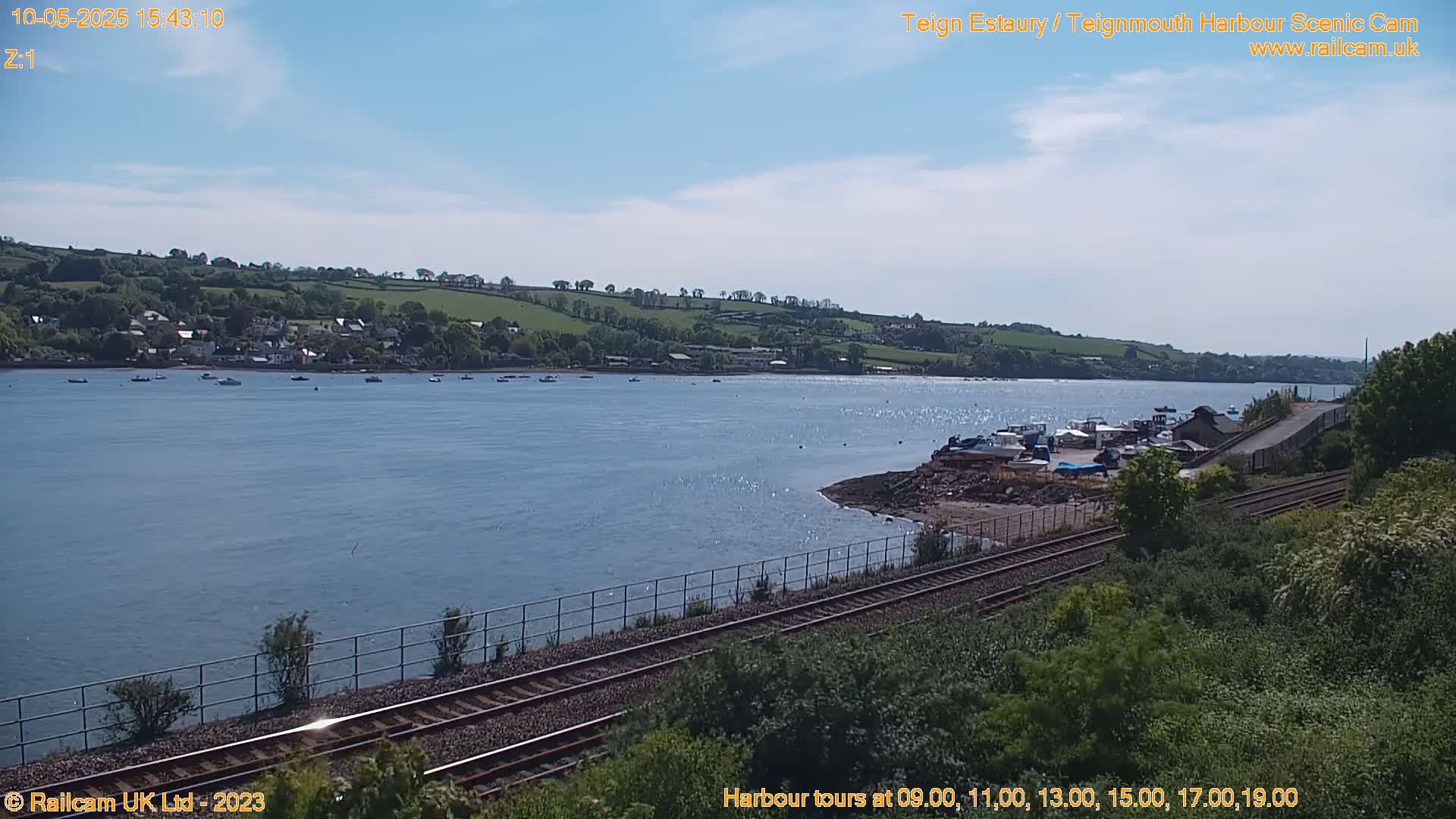 A sunny day shows a calm body of water with boats docked near a railway track, alongside a small town nestled on a hillside.