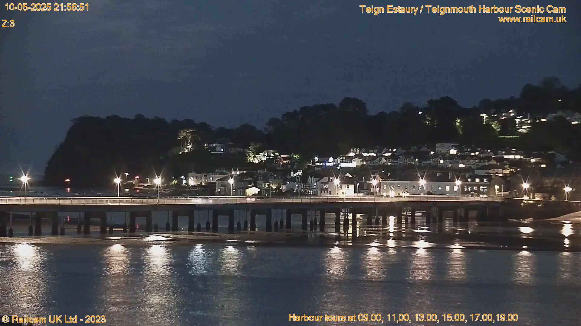 A nighttime view of a town's lit harbor and bridge reflected in calm water under a dark sky.