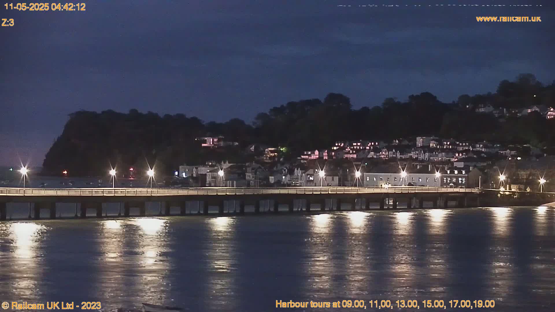 A long pier stretches across calm, dark water at night, illuminated by lights, toward a hilltop town silhouetted against a twilight sky.