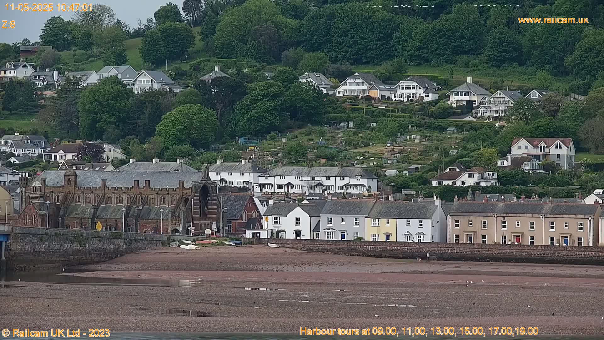 A coastal town with multi-story buildings and houses on a hillside, overlooking a wide, exposed sandy beach under an overcast sky.