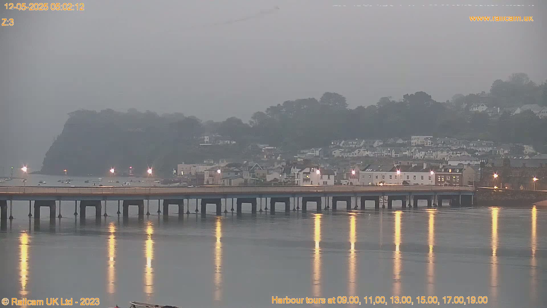 A long bridge spans a calm body of water reflecting lights on a foggy or overcast morning, with a town and hills visible in the background.