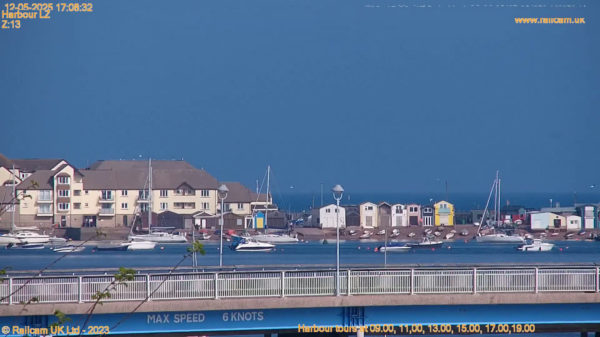 A harbor scene on a sunny day shows a calm body of water with boats and a building, a bridge with a speed limit sign, and colorful beach huts.