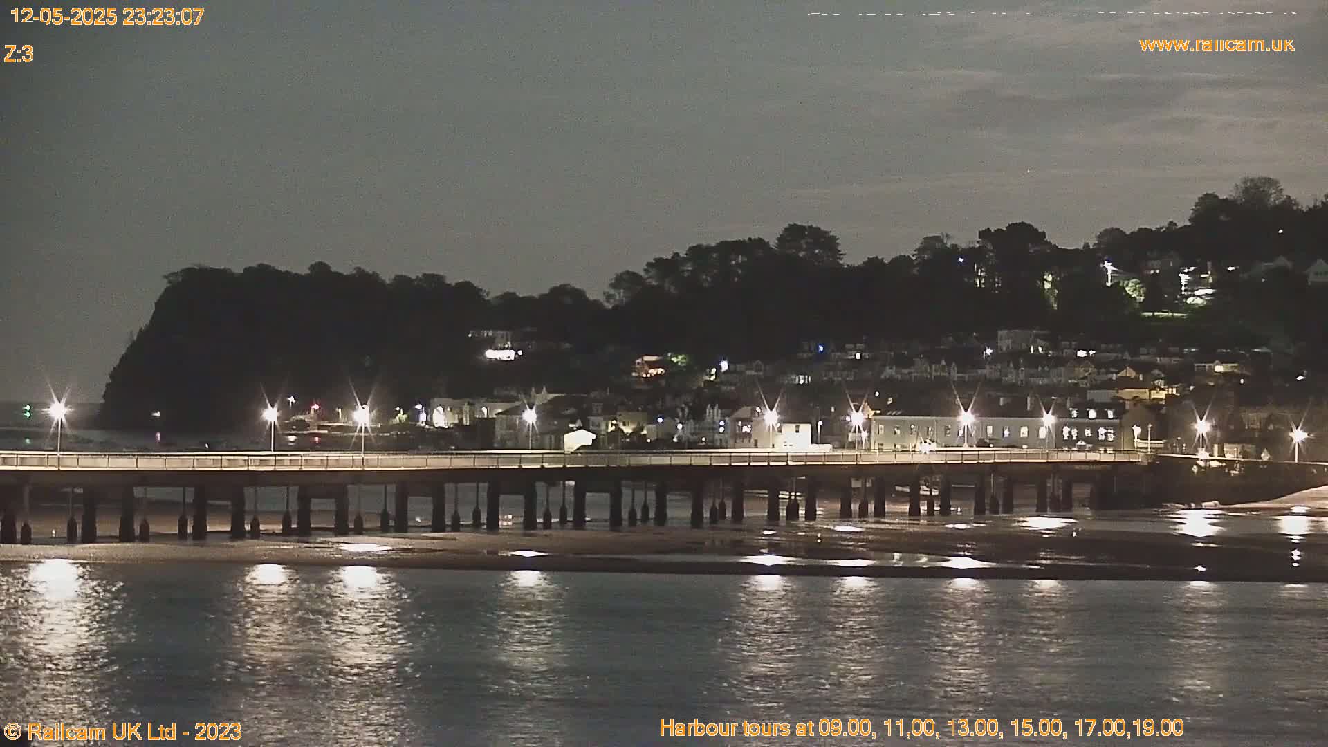 A long pier with lights extends over calm, dark water at night, with a hilltop town visible in the background under a mostly clear sky.
