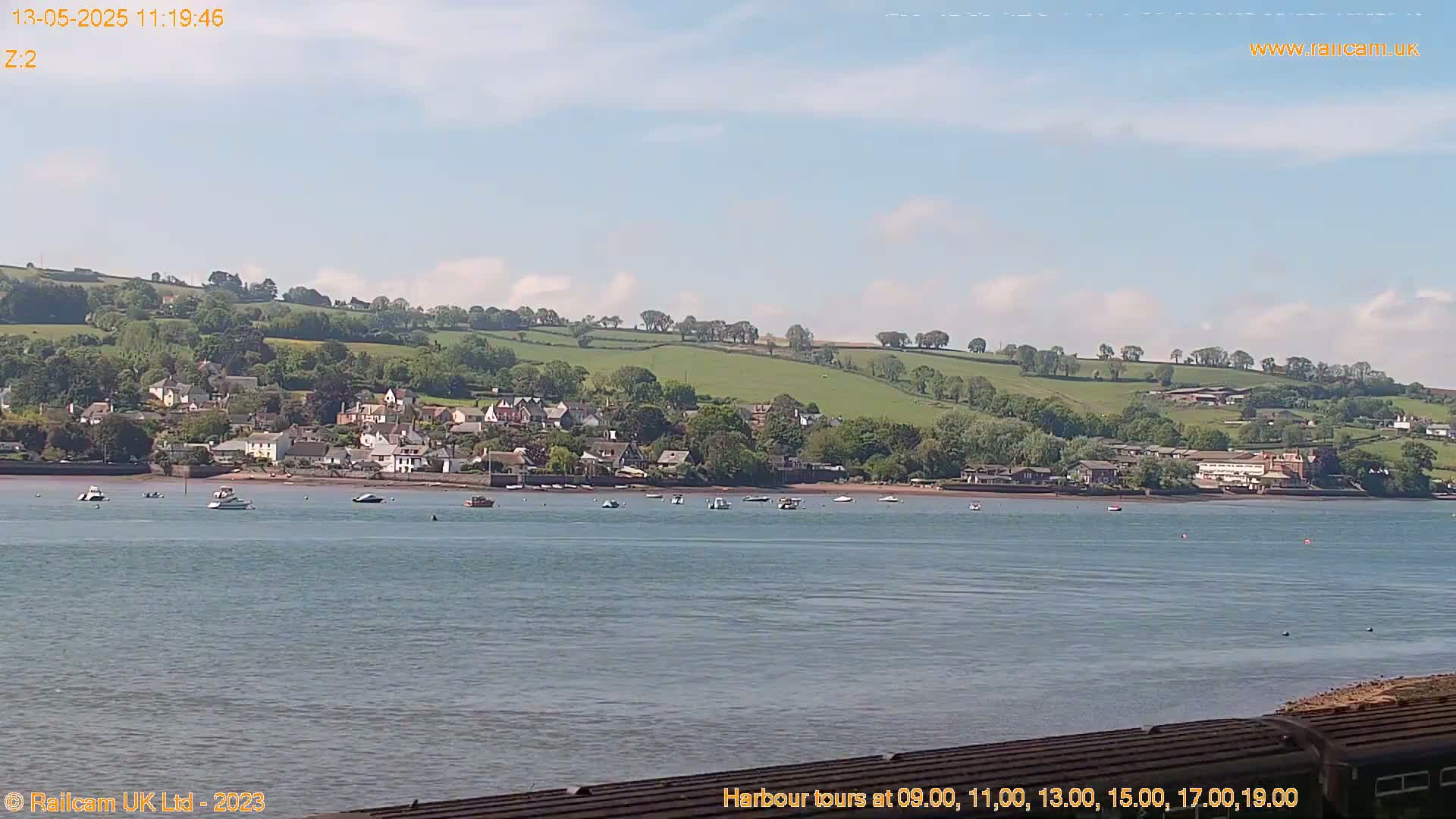 A calm body of water shows a small town nestled against a verdant hillside under a mostly sunny sky.