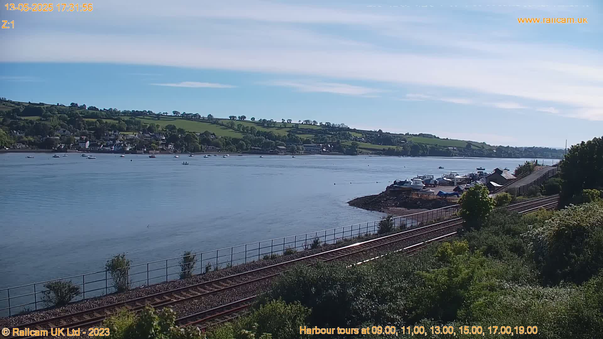 A calm body of water with several boats, a small town on a hillside in the background, and a railway line in the foreground under a clear, sunny sky.