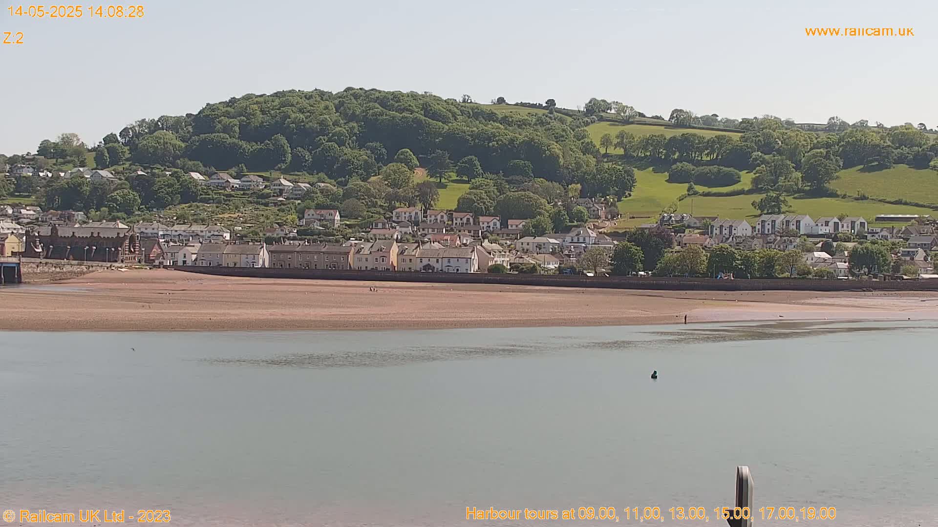 A coastal town nestled against a verdant hillside stretches along a sandy beach, with calm water in the foreground under a bright, sunny sky.