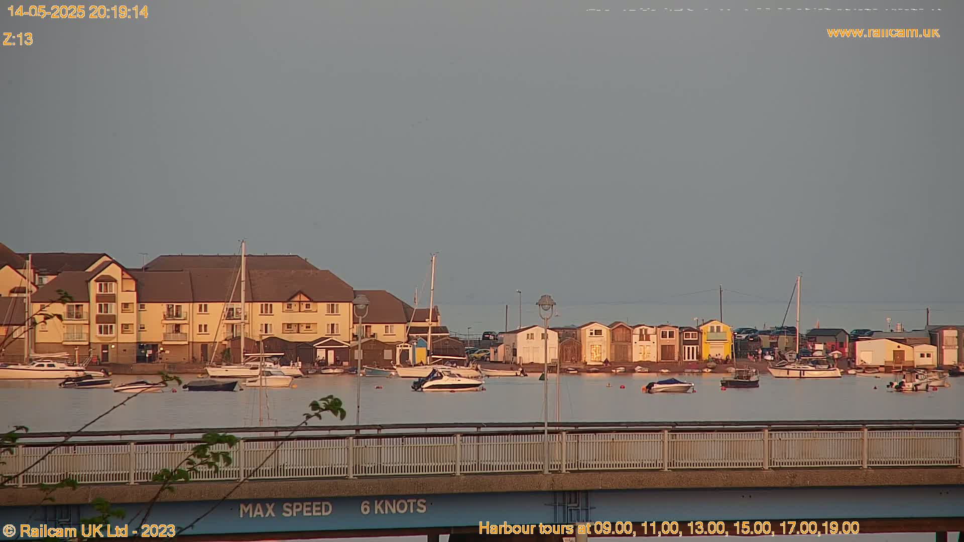 A calm harbor scene at dusk shows several boats moored near a row of buildings and small sheds, viewed from a bridge.
