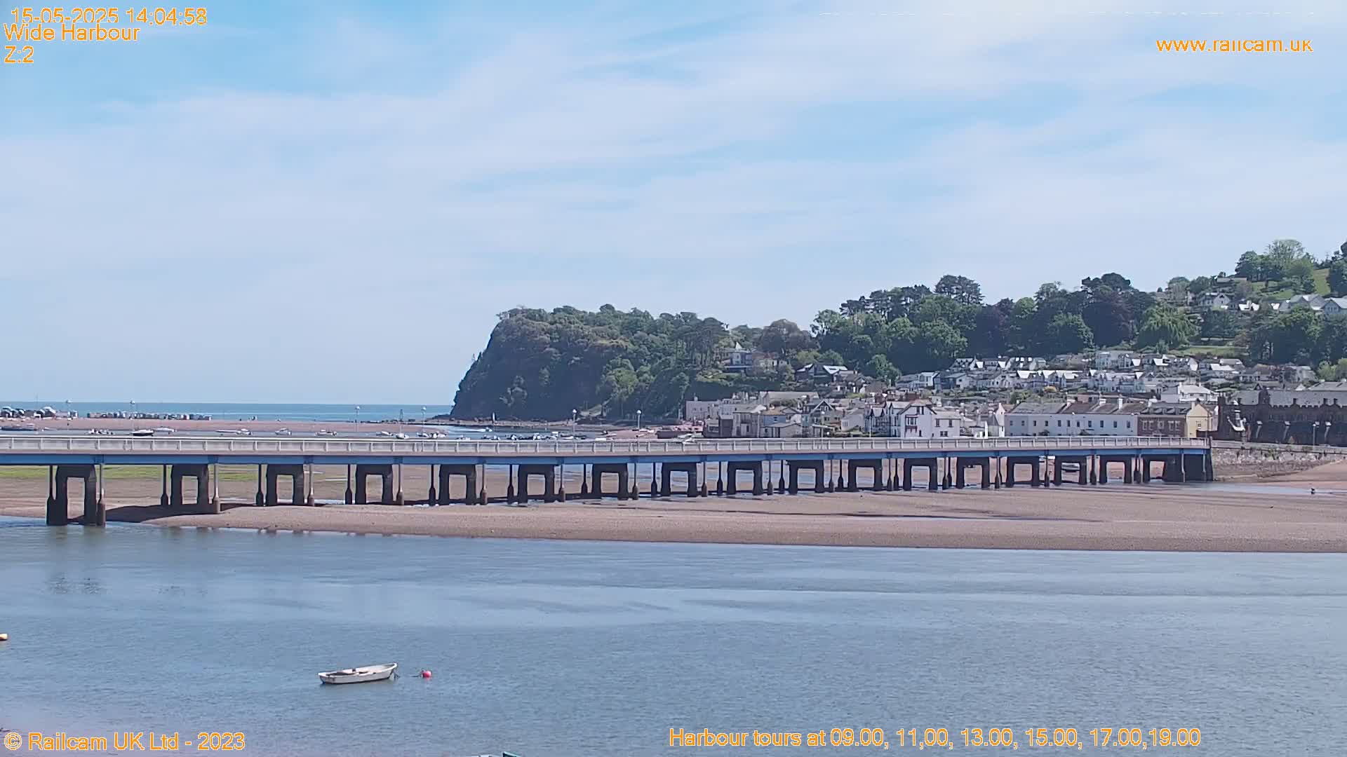 A long bridge stretches across a sandy expanse of water, leading to a town nestled against a verdant hillside under a clear, sunny sky.