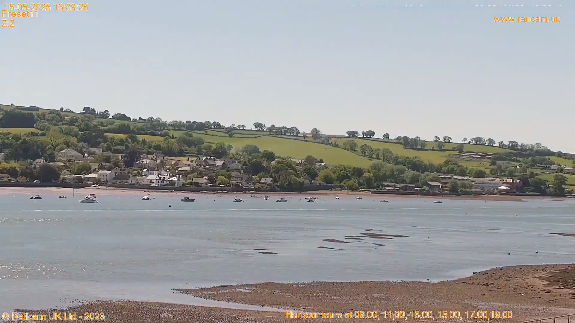 A calm, sunny day reveals a coastal town nestled along a river, with several boats visible in the water and a gently sloping hillside in the background.