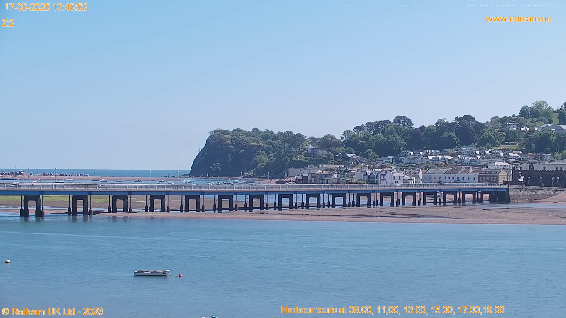 A long bridge spans a calm body of water under a clear blue sky, with a small town and a verdant hill visible in the background.