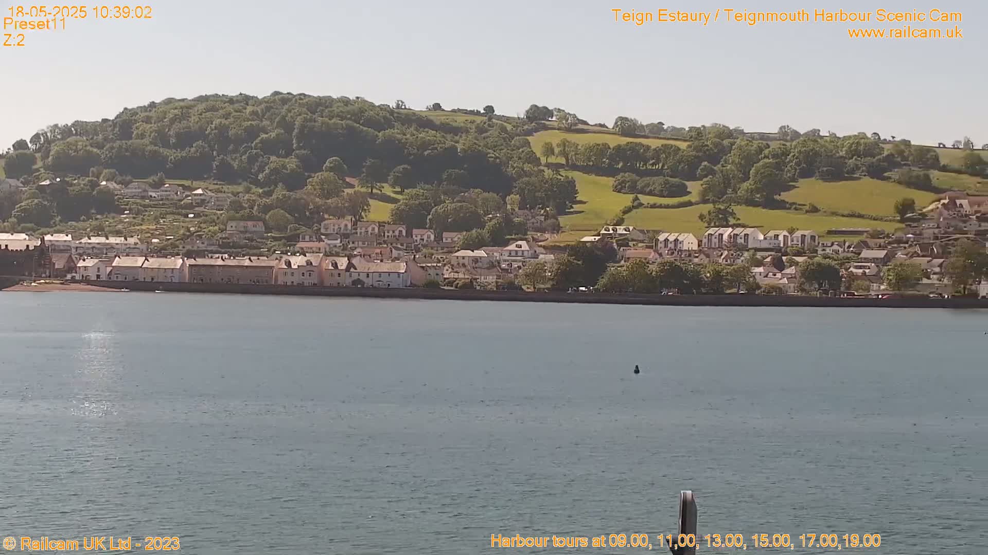 A seaside town sits below a verdant hill under a bright, sunny sky, with calm water in the foreground.