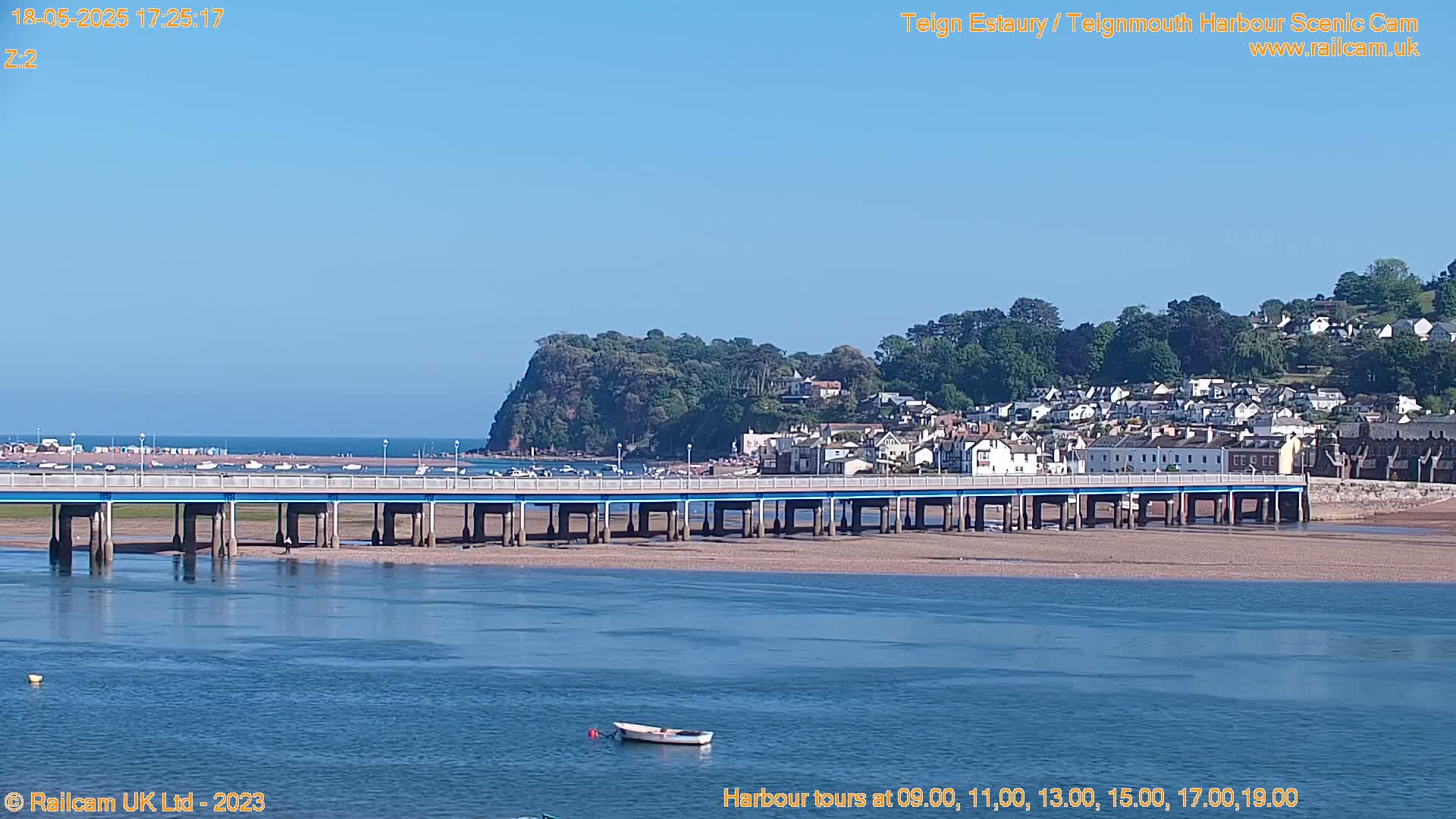 A long bridge spans a calm, blue waterway, with a small boat visible in the foreground,  under a clear blue sky; a town and green hill are visible in the background.