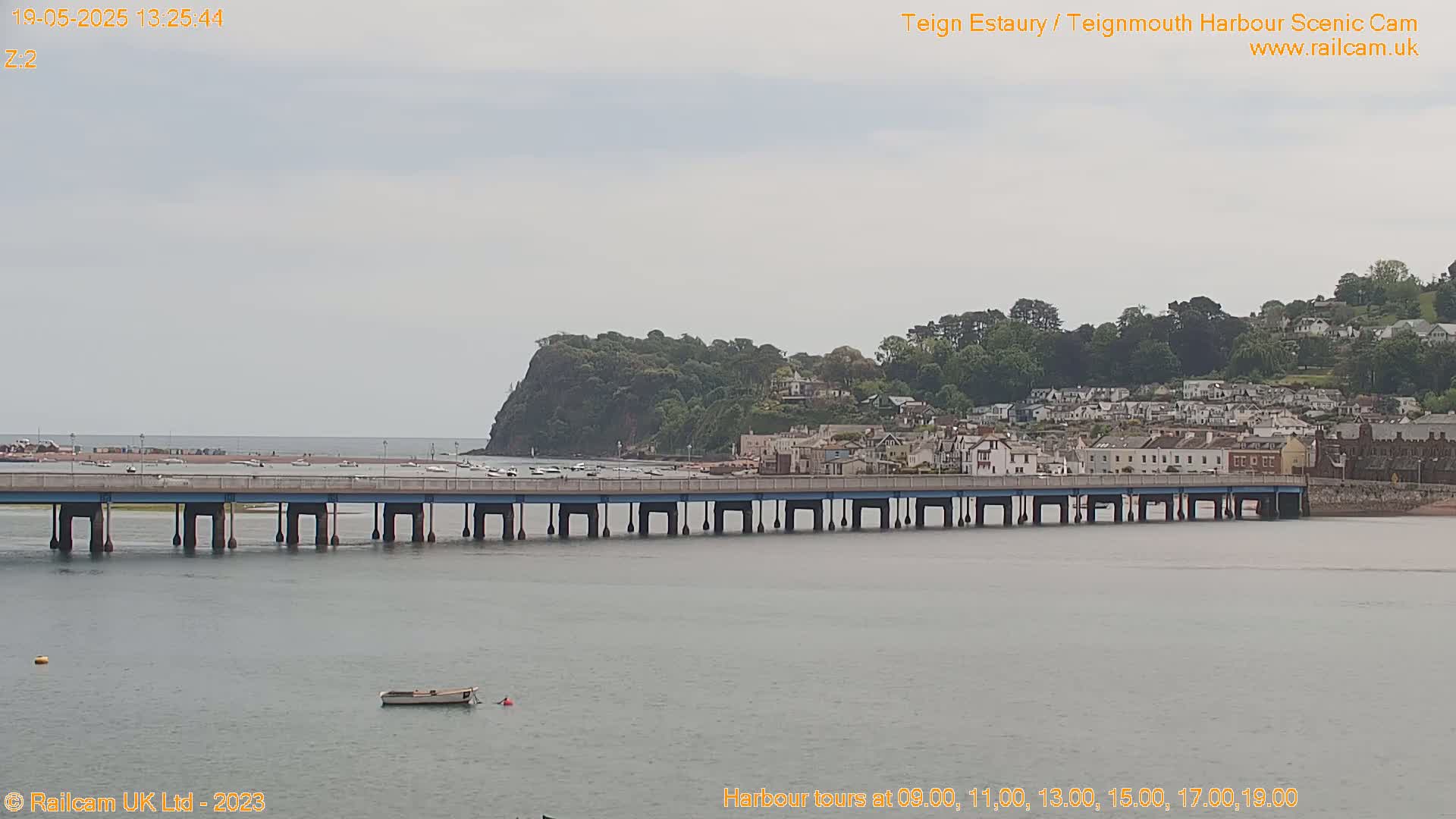 A long bridge stretches across calm, gray water toward a town nestled against a verdant hillside under an overcast sky.