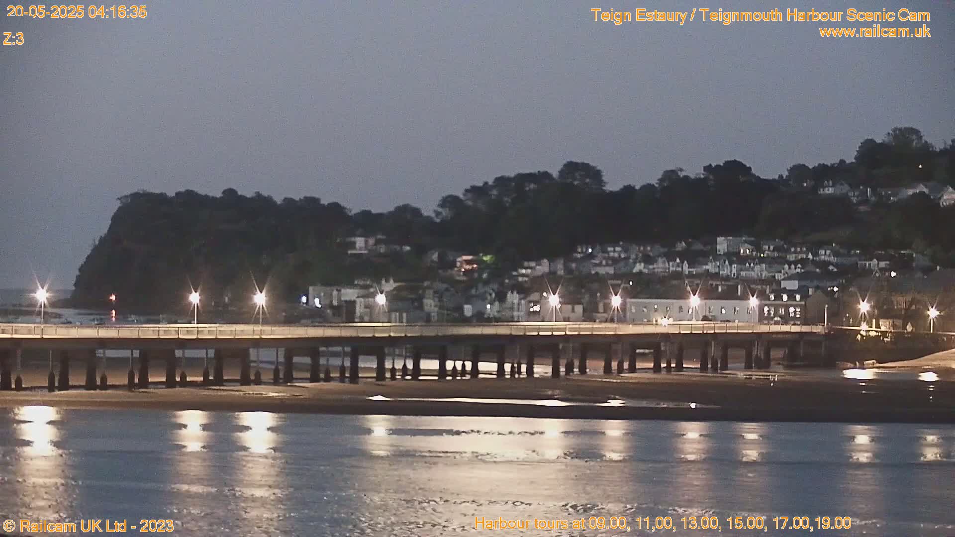 A long pier with lights extends into a calm body of water under a twilight sky, with a town visible on the far shore.