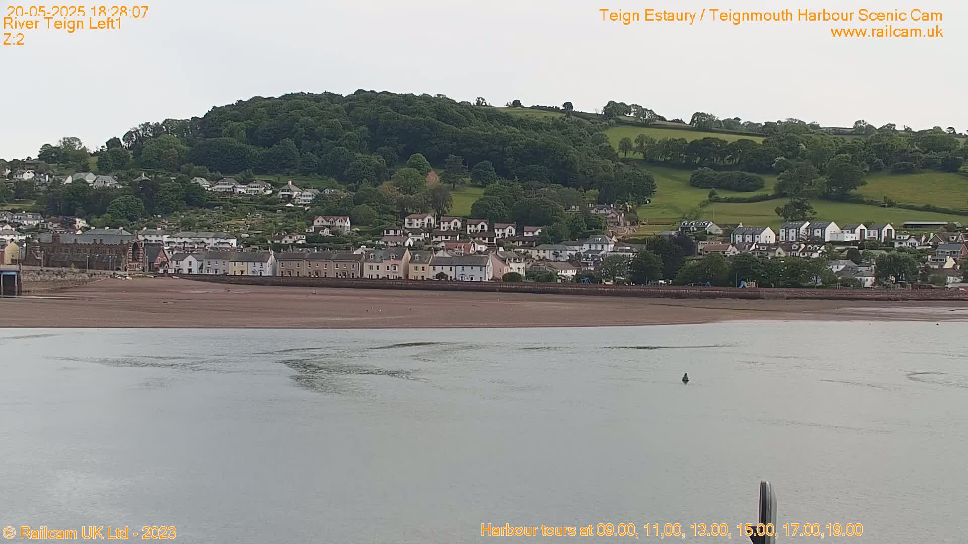 A coastal town sits at the base of a verdant hill beside a calm body of water under an overcast sky.