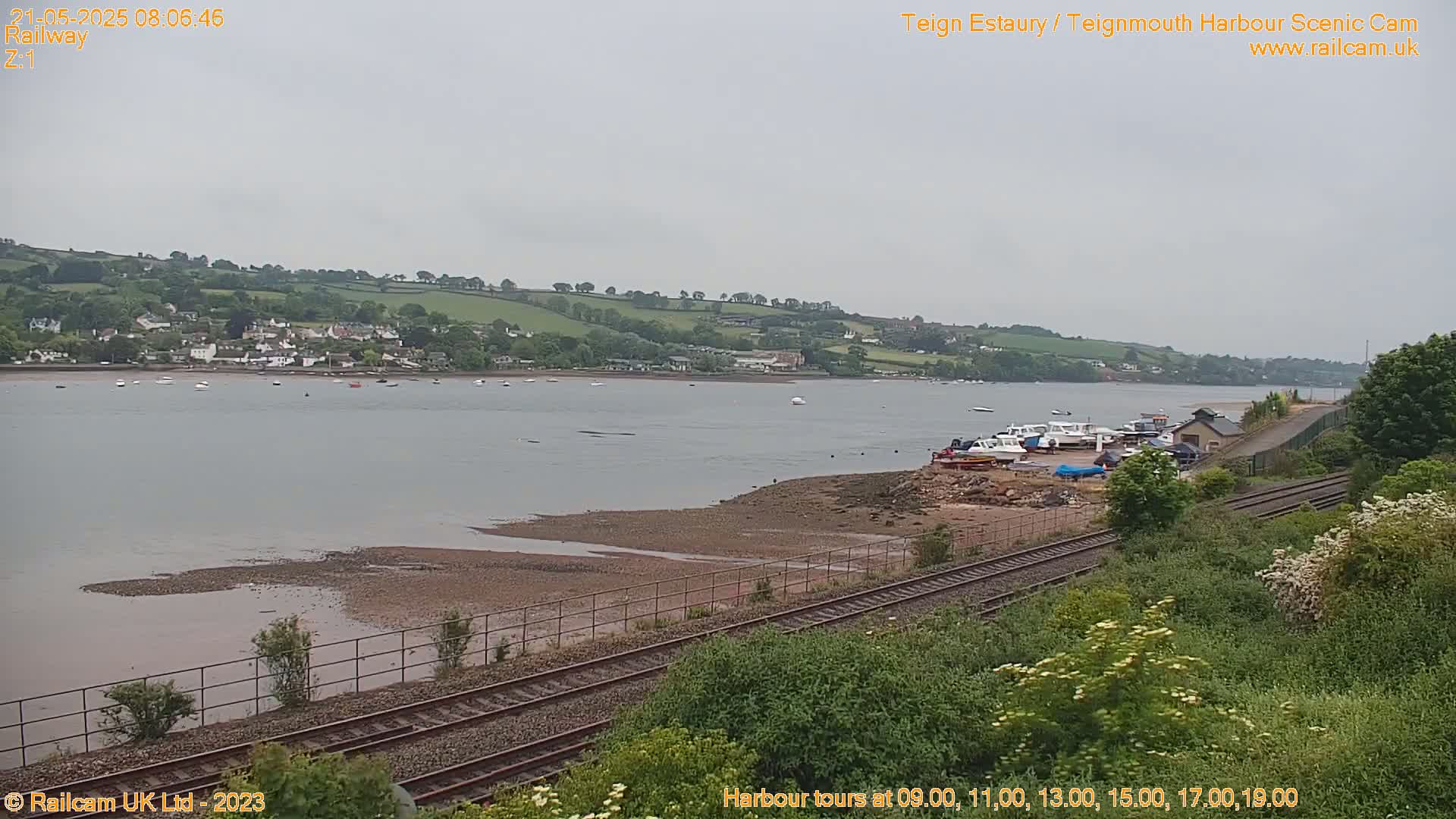 A calm, overcast day shows a view of a river estuary with several boats docked near a railway line and a small town visible on the opposite shore.