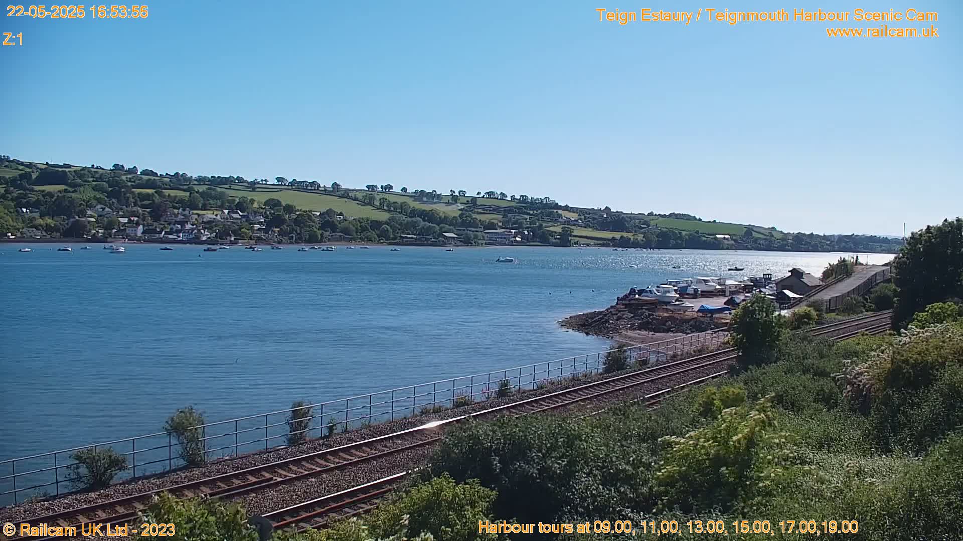 A calm, sunny day overlooks a body of water with several boats and a small town on the far shore, next to a railway line.