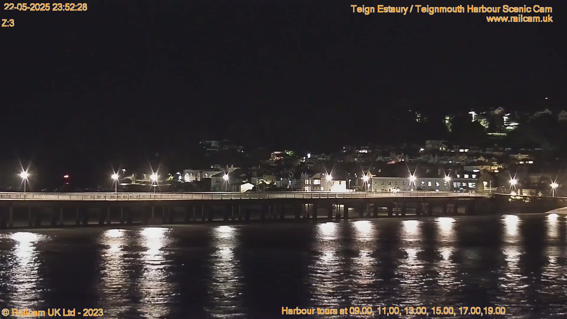A long pier at night is illuminated by lights, reflecting in calm water, with a town visible in the background under a dark sky.