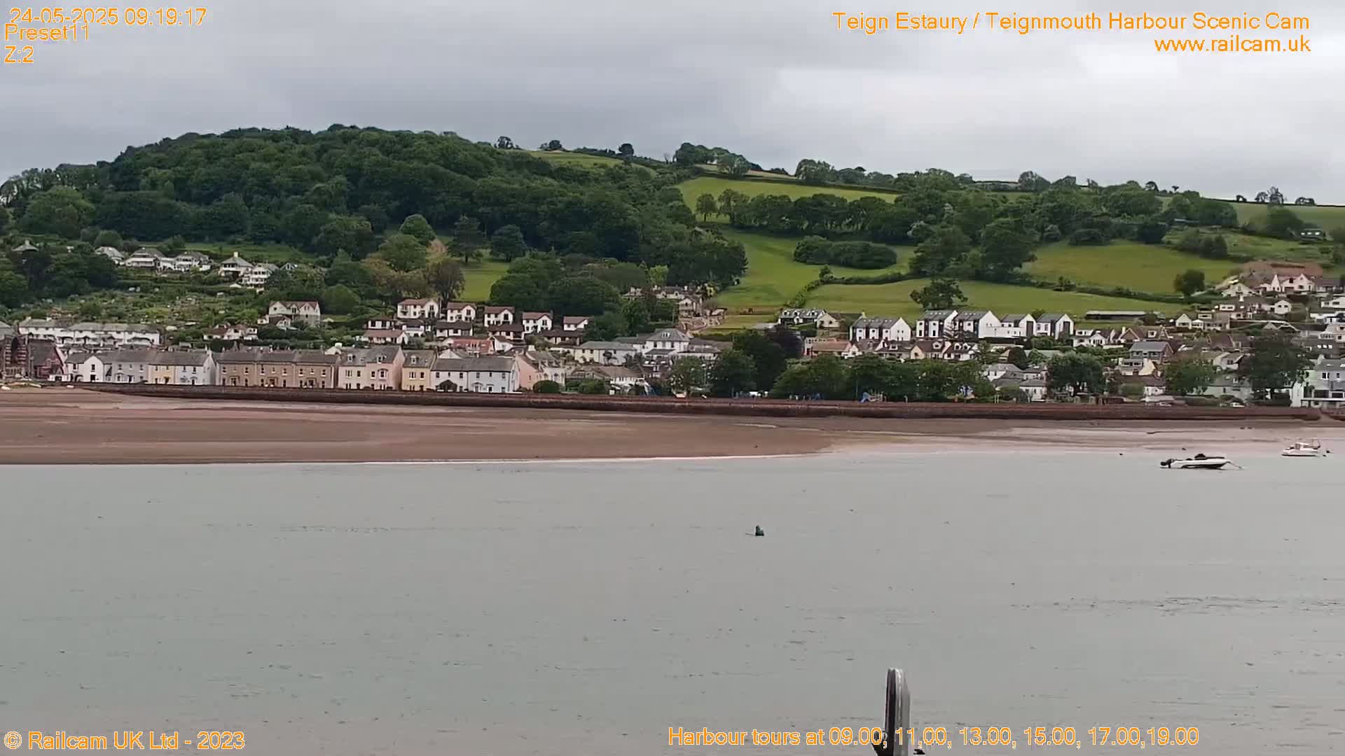 A coastal town nestled below a green hill is viewed from across a calm body of water under an overcast sky.