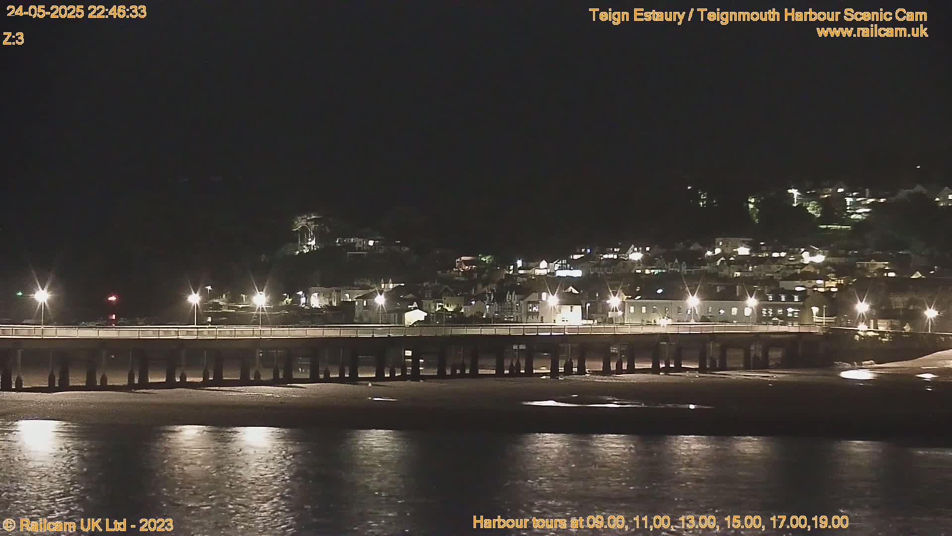 A long pier stretches across a dark, calm body of water at night, with a town's lights illuminating the hillside beyond.