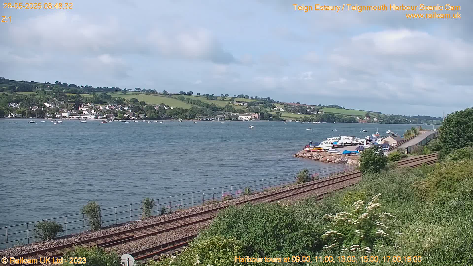A wide shot of a calm body of water with boats and a shoreline featuring a train track, under a partly cloudy sky.